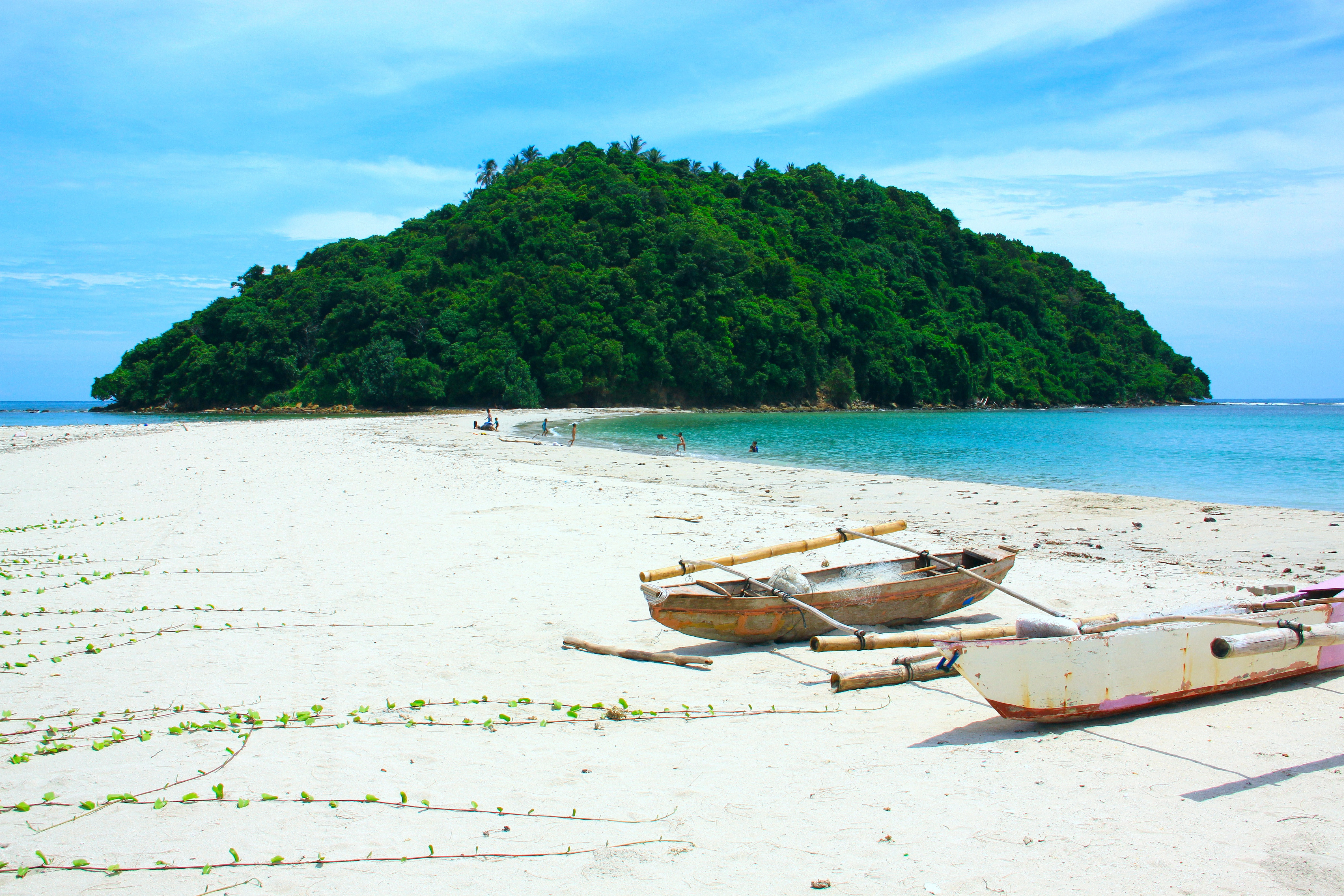 Tropical beach with white sand, traditional wooden boats, and a lush green island in the background, under a clear blue sky