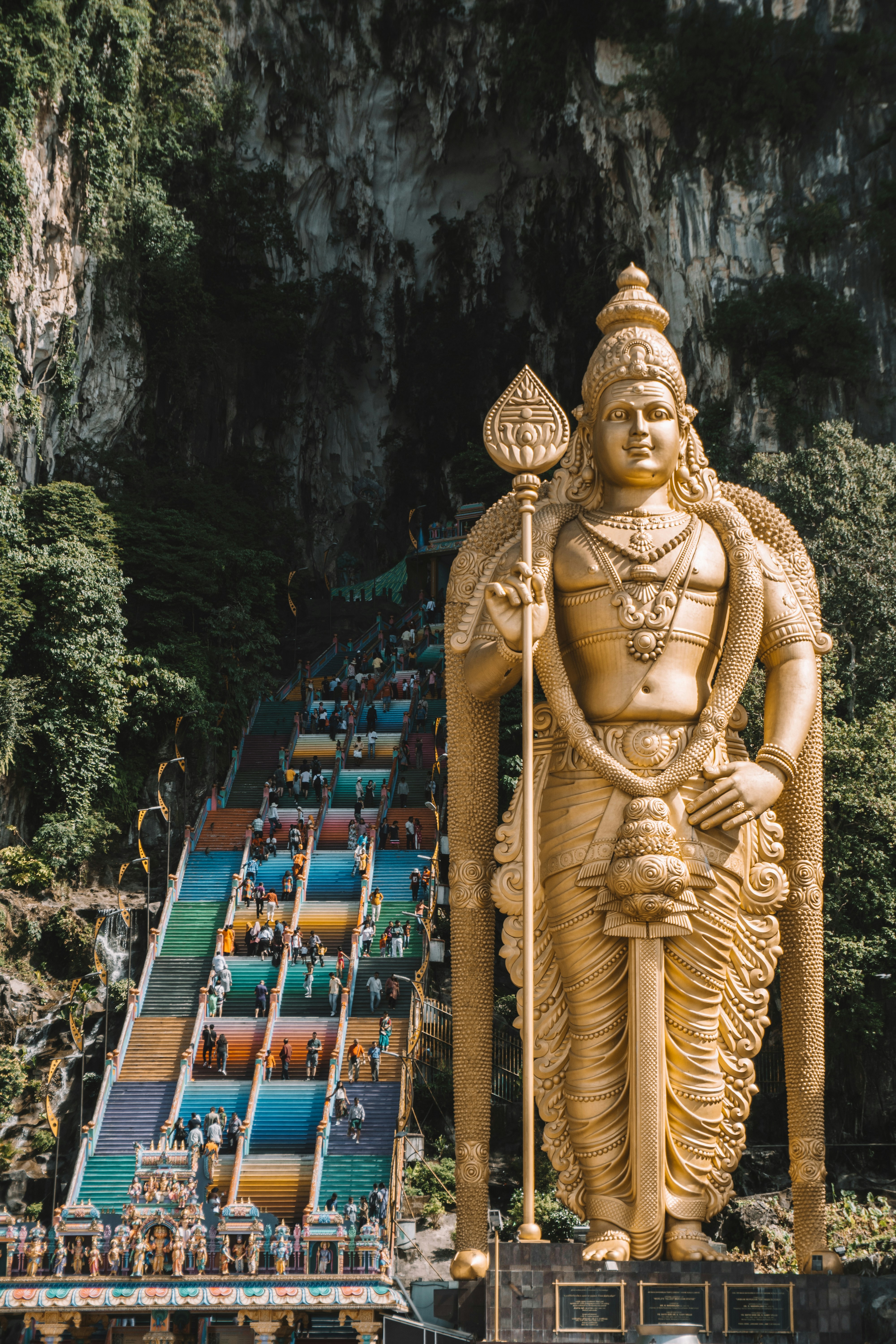 Gyllene staty av Lord Murugan vid ingången till Batu-grottorna i Malaysia, med en färgglad trappa som leder till tempelgrottan, omgivet av frodiga gröna klippor