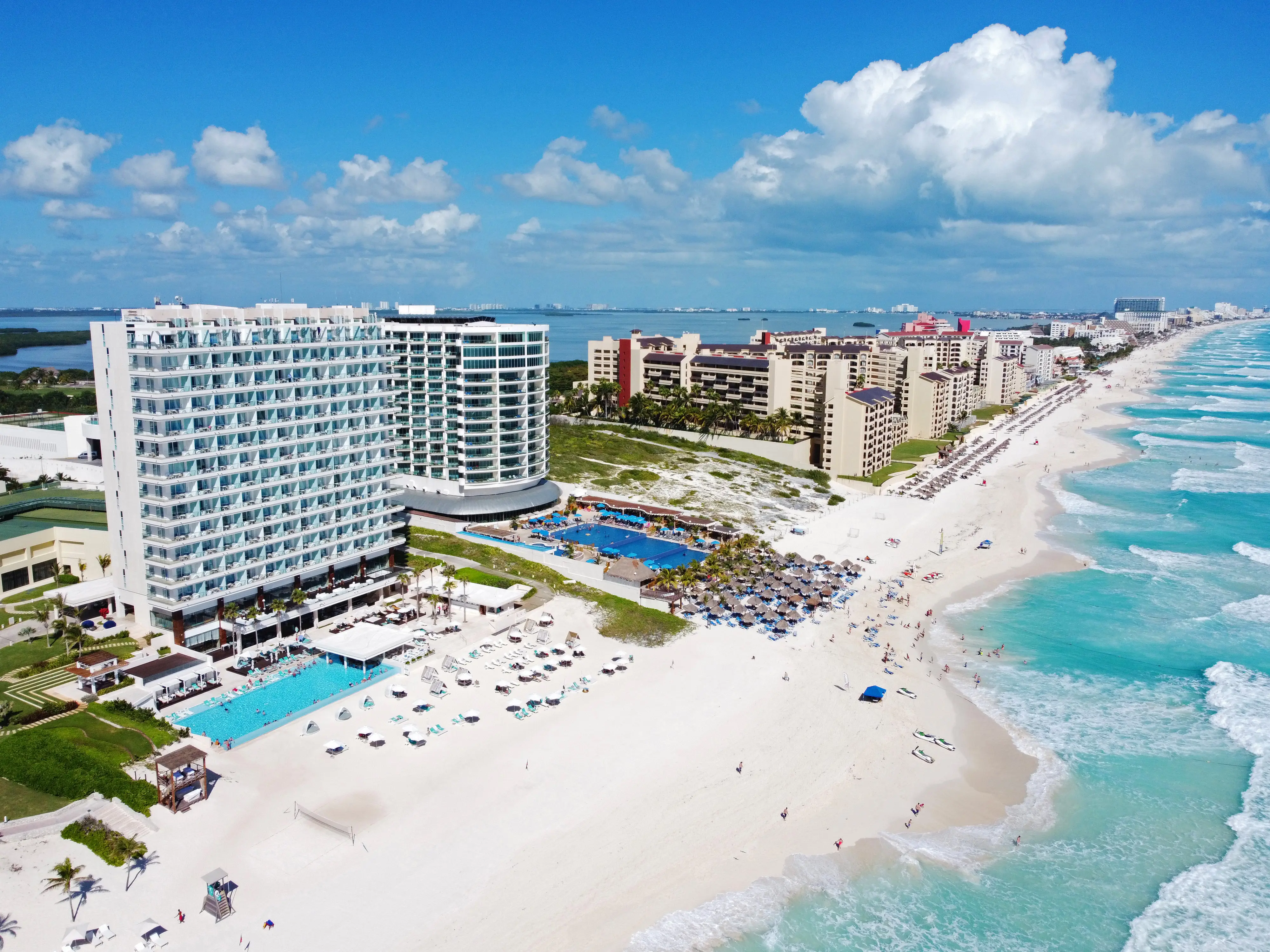 Aerial view of a beachfront resort with modern high-rise buildings, a large swimming pool, and white sandy beach in Cancun, Mexico. The turquoise ocean waves gently lap against the shore under a partly cloudy sky.