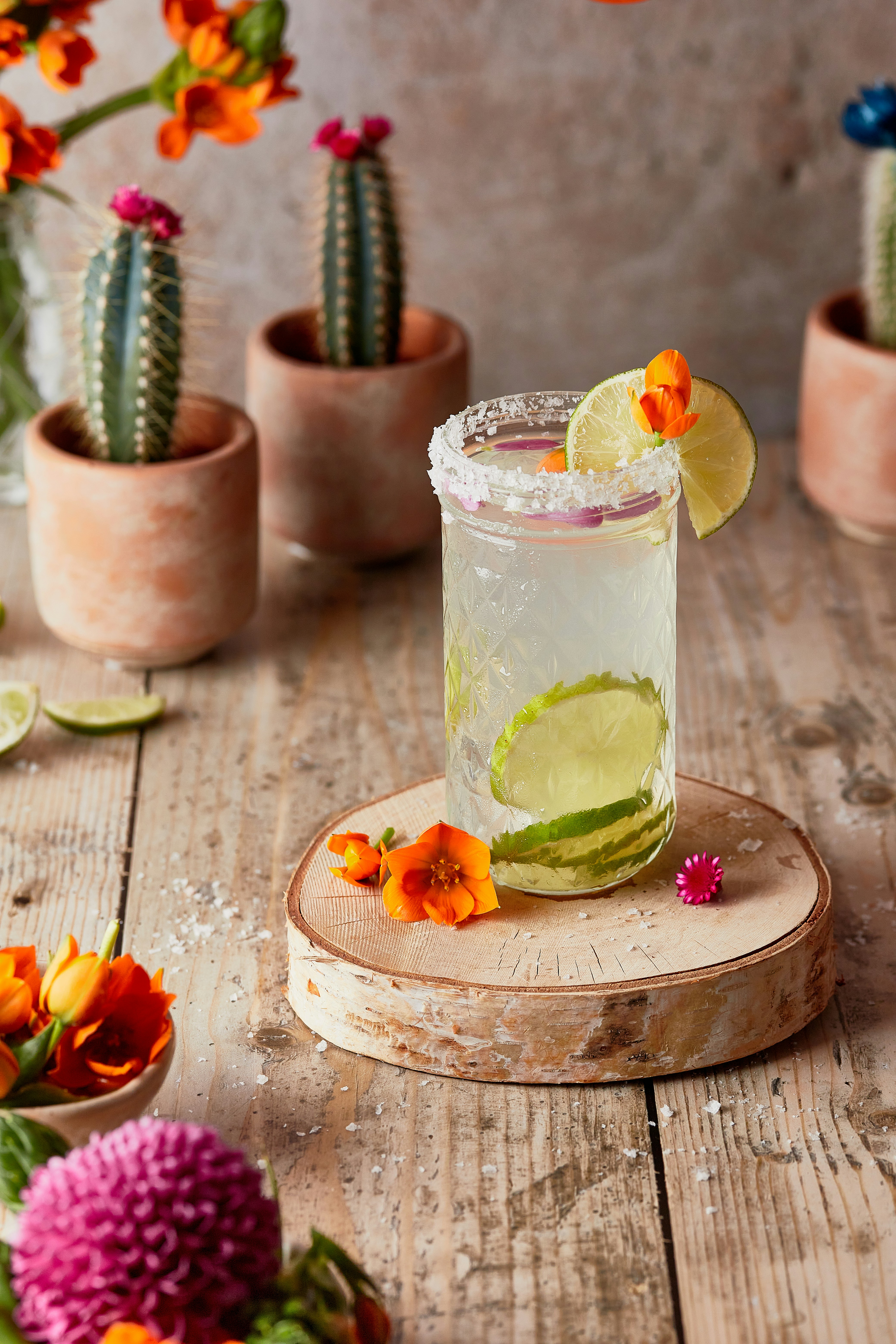 Uppfriskande limecocktail garnerad med en skiva lime och orange blommor, serverad i ett glas med saltad kant på ett rustikt träbord, omgiven av krukväxter och livfulla blommor