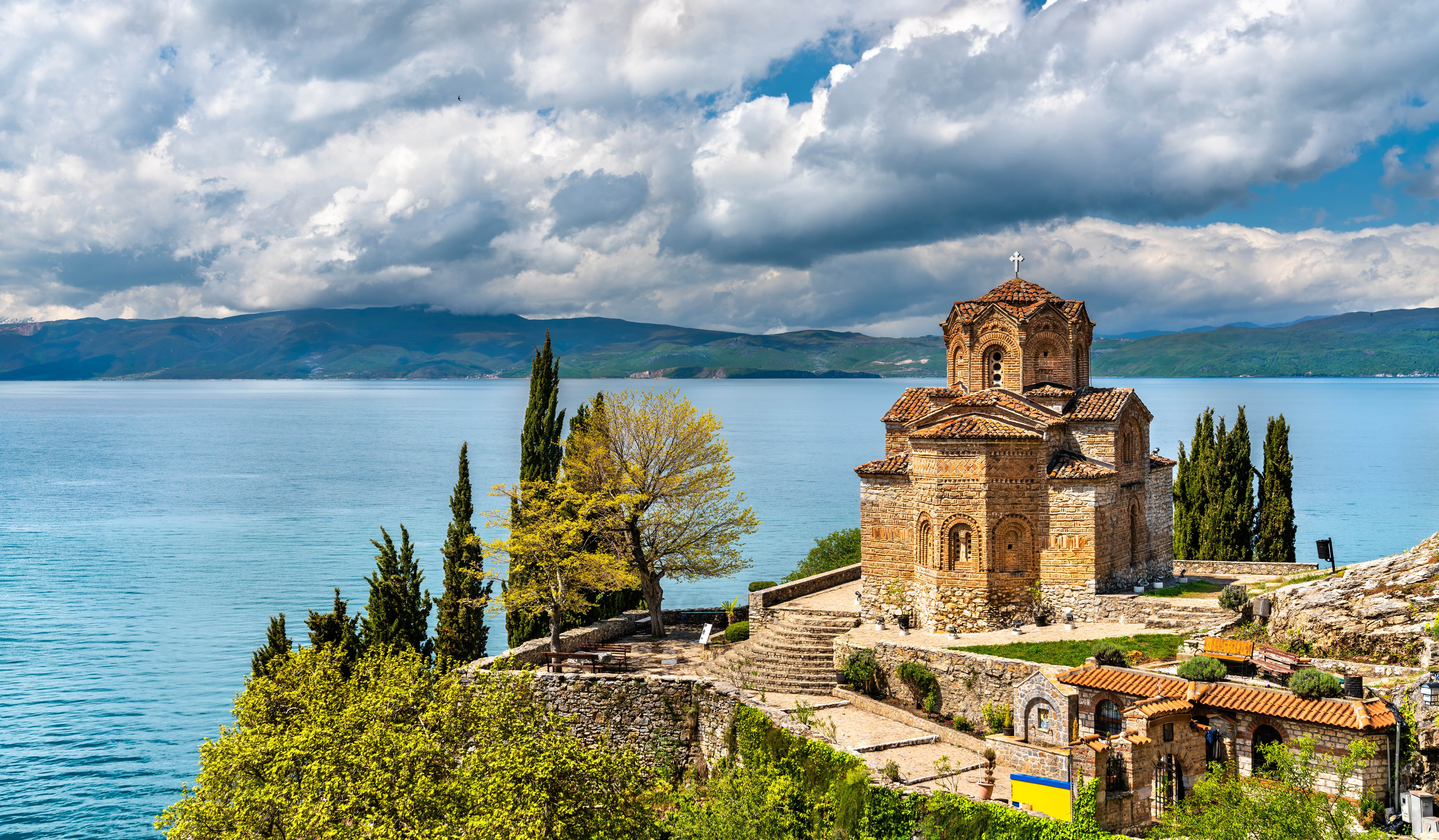 Vanha kivikirkko kauniilla rannalla pilvisenä päivänä, taustalla vuoristo ja sininen järvi.