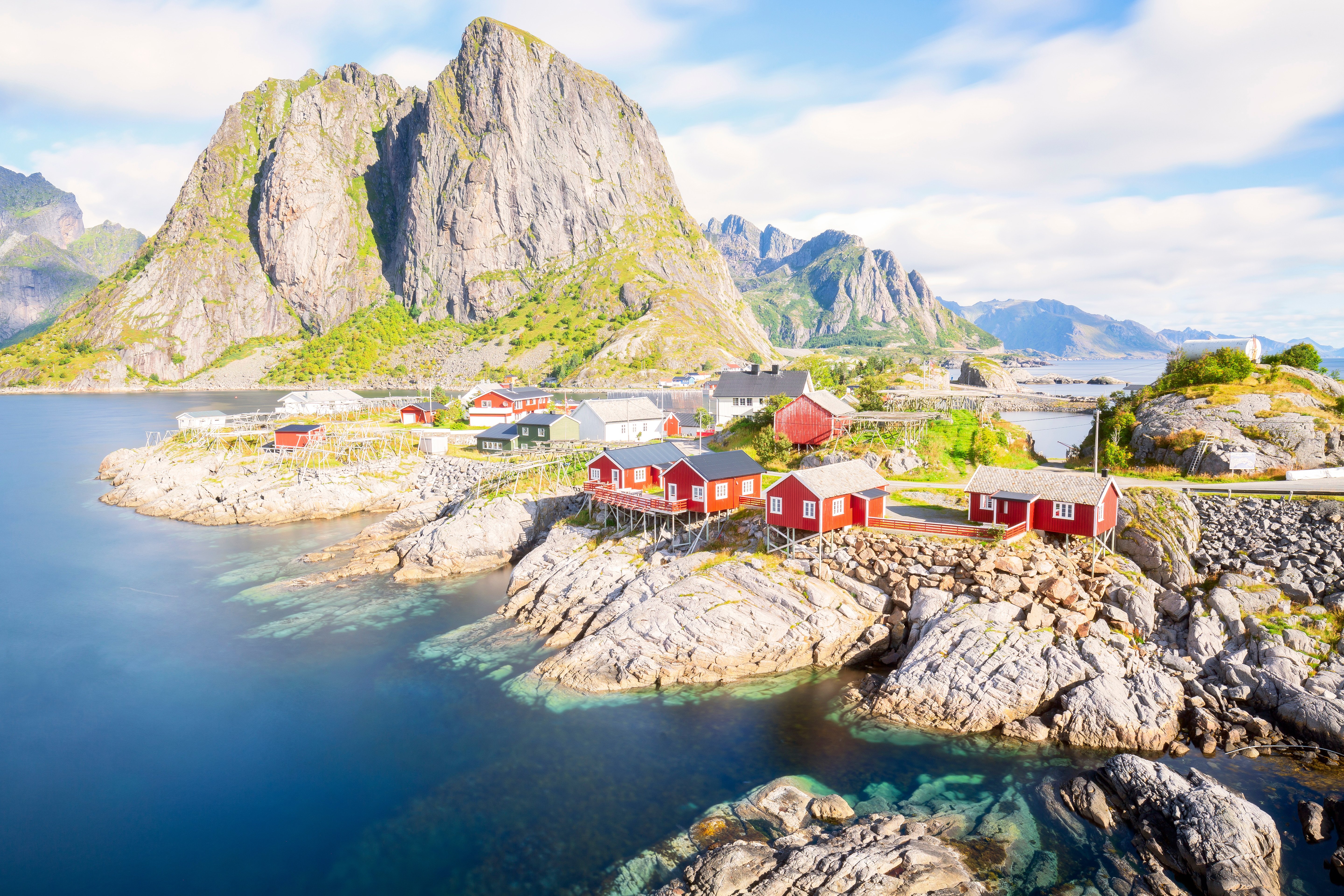 Punaiset kalastajamökit kauniilla rannalla, taustalla vihreät vuoret ja sininen taivas, Lofoten, Norja