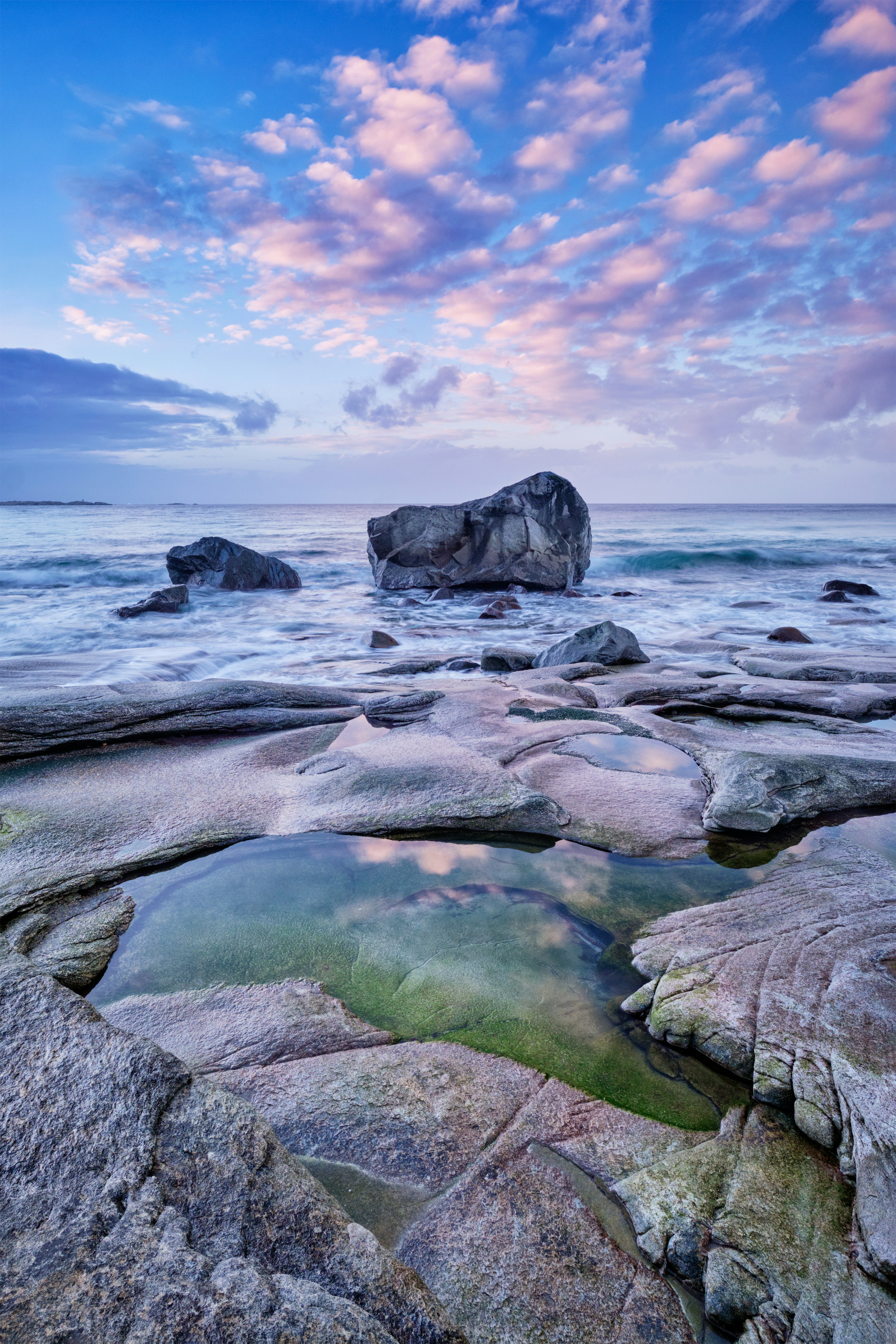 Ranta kivillä, taustalla sininen meri ja vaaleanpunaisia pilviä taivaalla