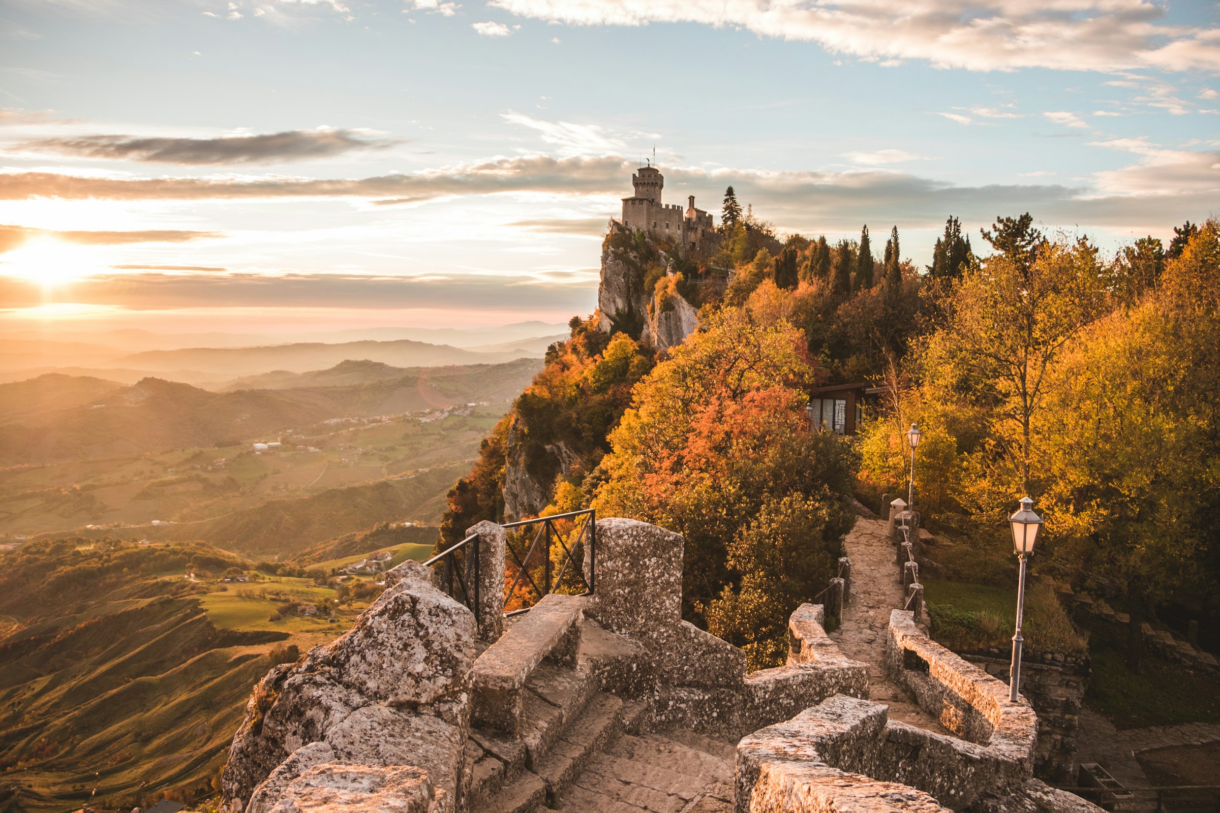 Sunset over a medieval castle on a cliff surrounded by autumn colors in San Marino, with mountainous views in the background.