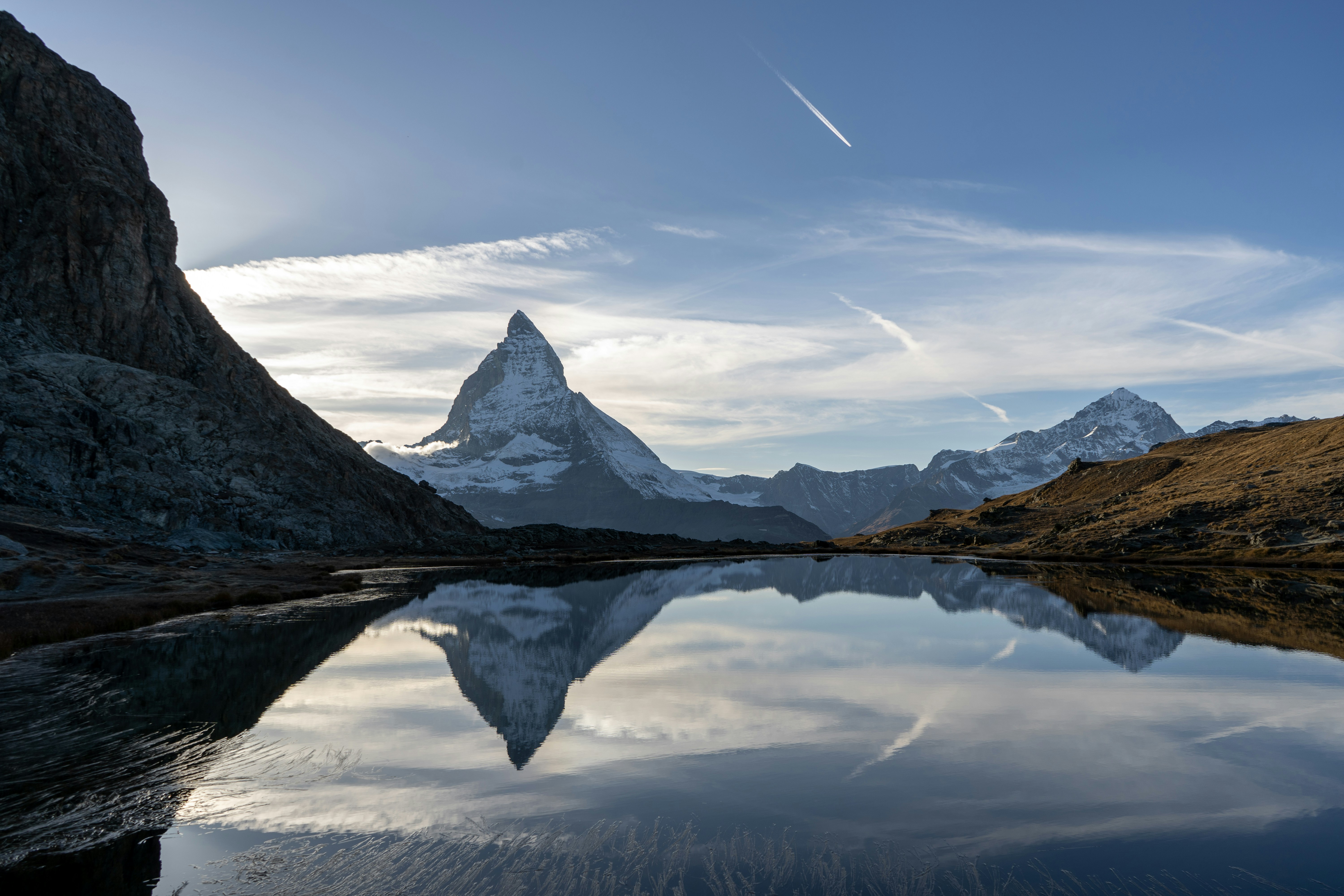 Snötäckt Matterhorn-berg reflekteras i en lugn sjö under en klarblå himmel med moln, omgiven av robust terräng