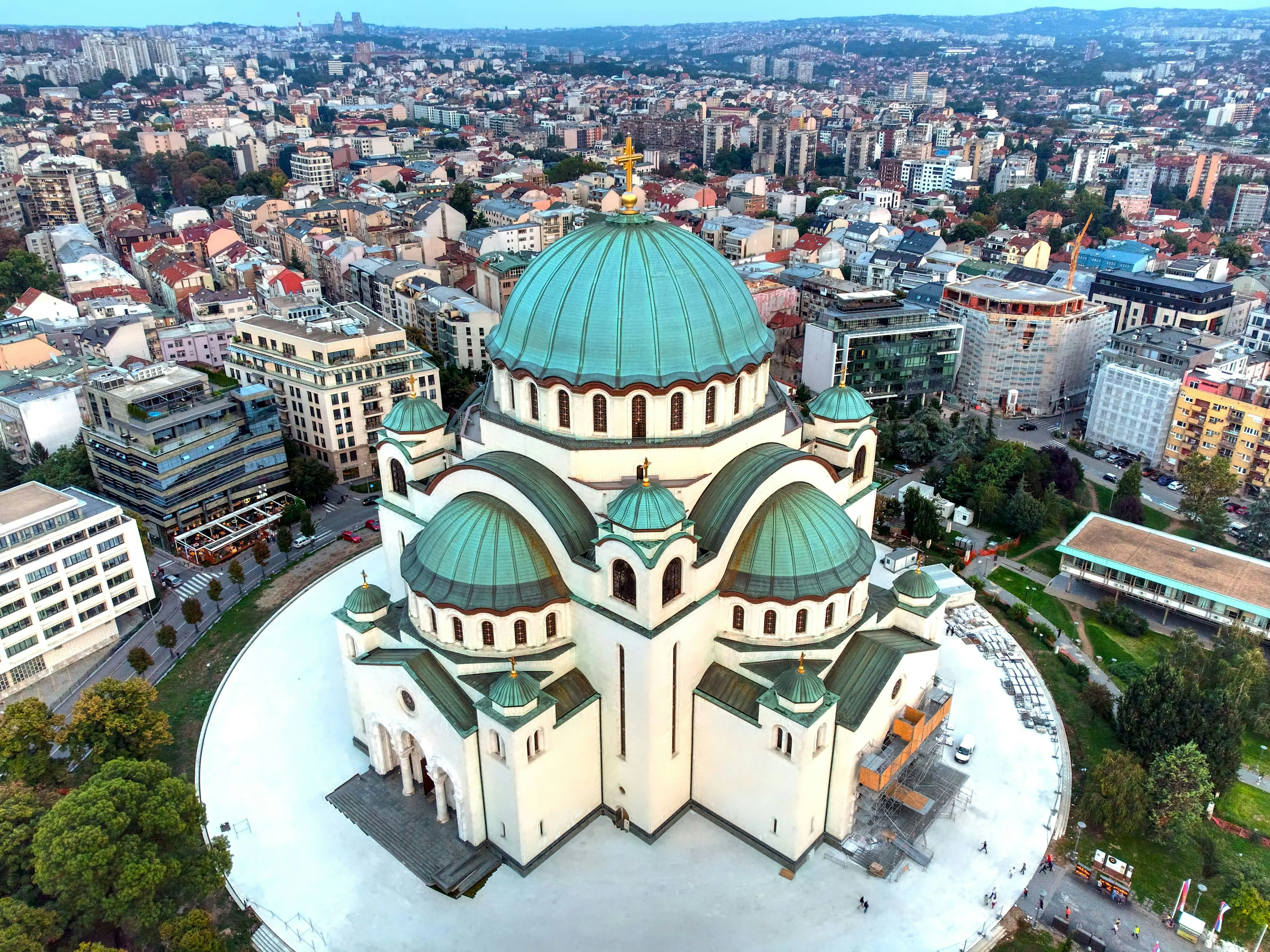 Luftvy över Sankt Sava-kyrkan i Belgrad med stadens silhuett i bakgrunden. Katedralens karakteristiska gröna kupol står i kontrast mot den tätt bebyggda omgivningen