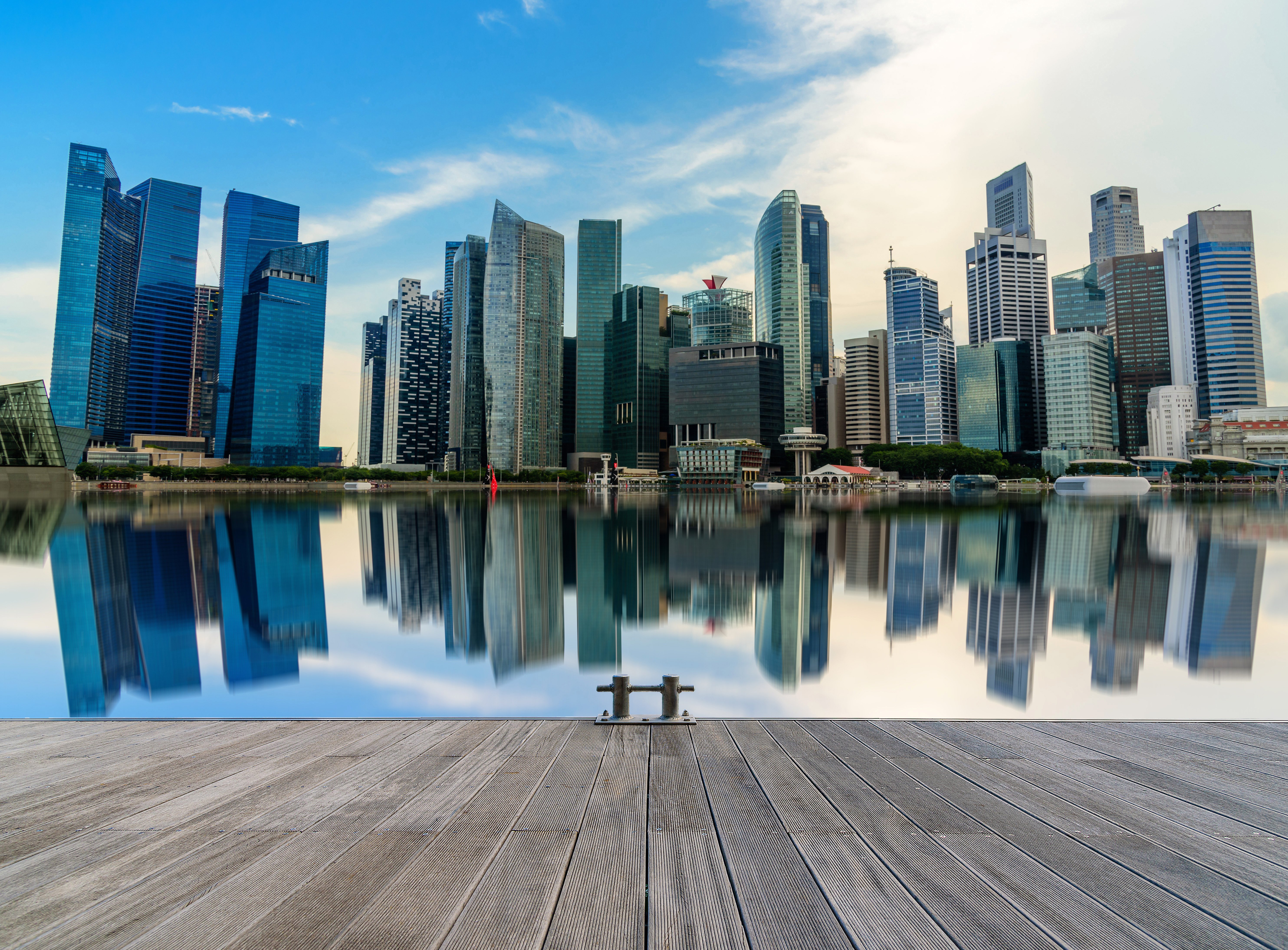 Modernin Singaporen siluetti kohoavine pilvenpiirtäjineen, jotka heijastuvat veteen, katsottuna puuterassilta kirkkaana päivänä