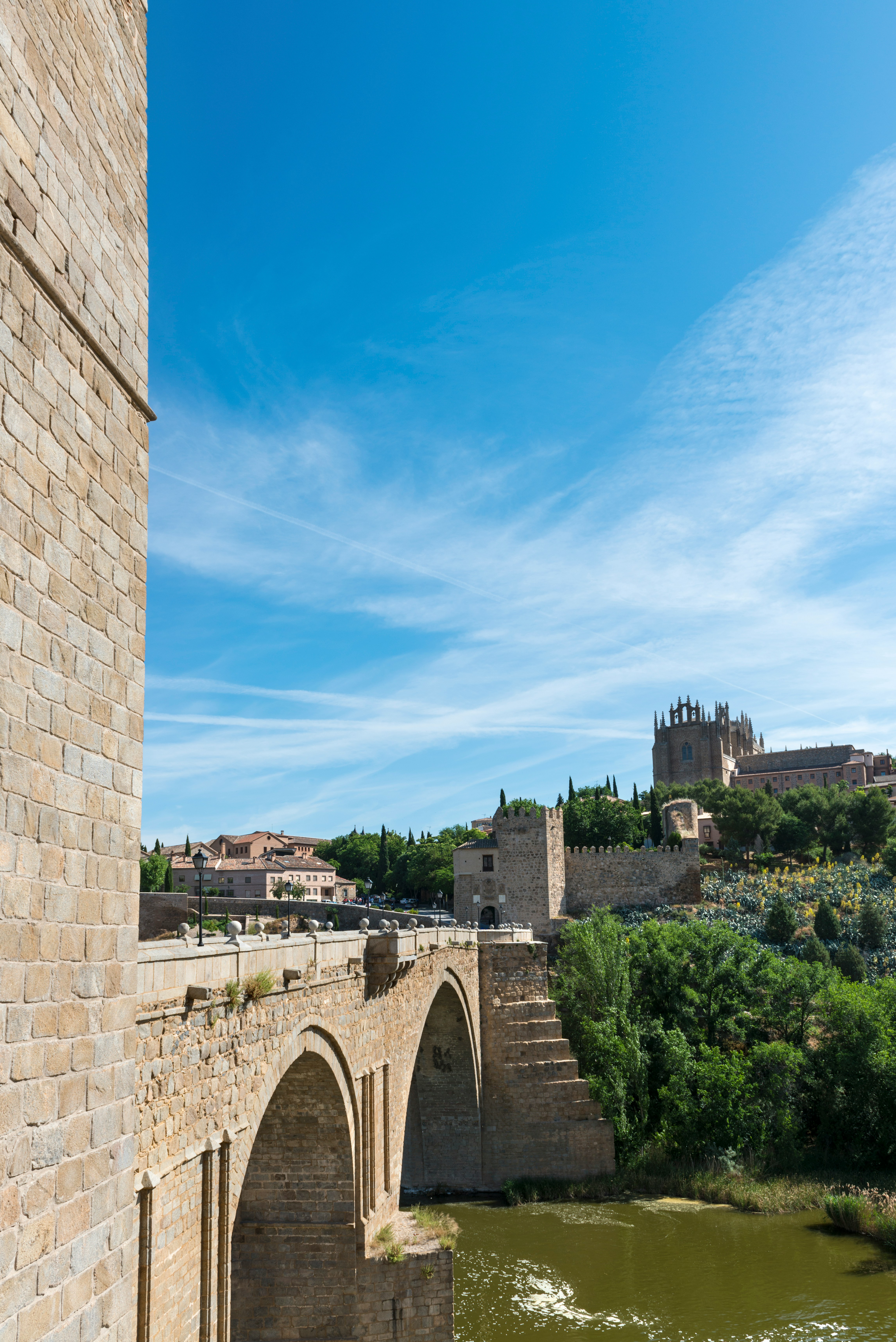 Historisk stenbro över floden i Toledo, Spanien, med medeltida arkitektur och frodig grönska under en klarblå himmel