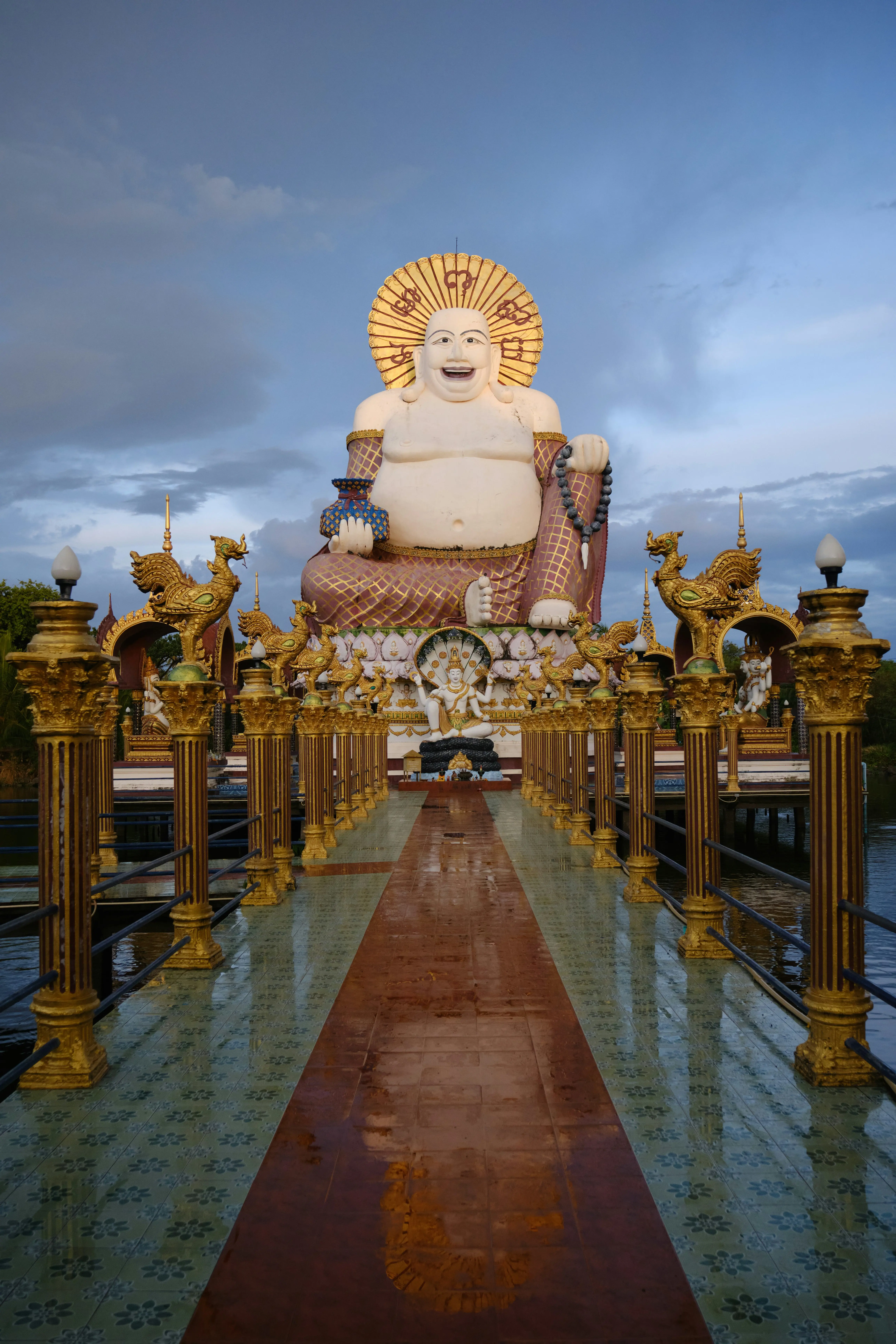 Suuri valkoinen Buddha-patsas kultaisin koristeluin Wat Plai Laem -temppelissä Koh Samuilla, Thaimaassa, nähtynä koristellulta kävelytieltä, jolla on koristeellisia pylväitä auringonlaskun aikaan
