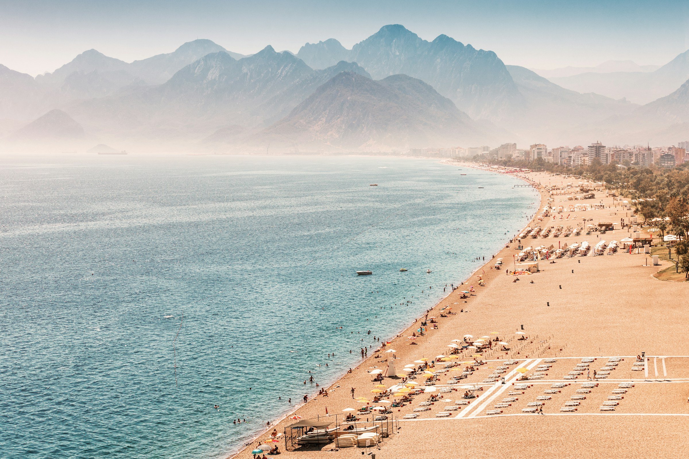 Sunny beach in Antalya, Turkey, with umbrellas and lounge chairs along the shoreline, clear blue water, and distant mountains.