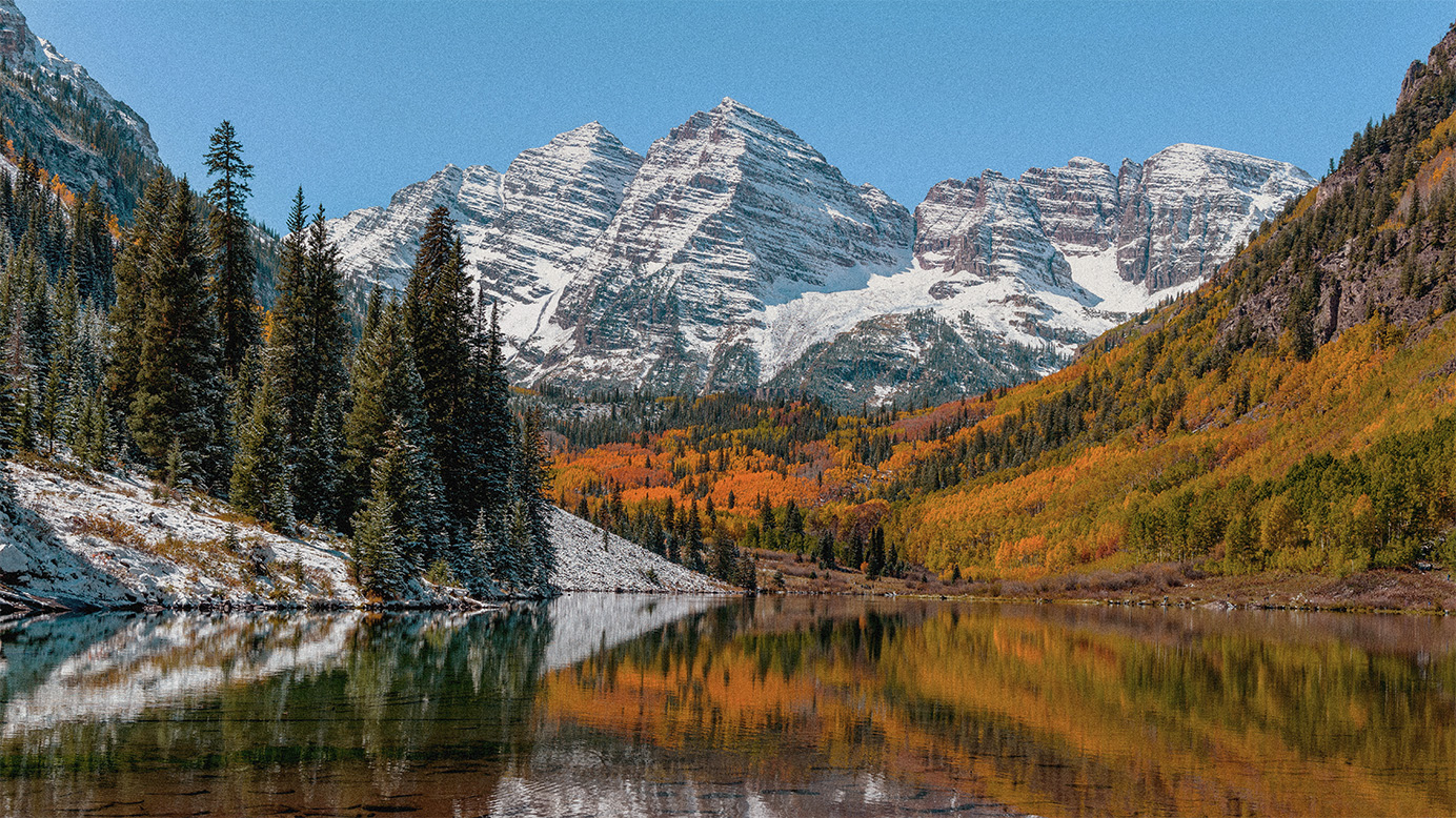 Snow-capped mountain peaks with autumn-colored trees and a mirror-like lake in the foreground in a scenic terrain.