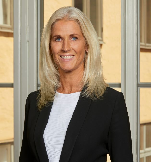 Vaaleahiuksinen nainen mustassa takissa seisoo ikkunan edessä.