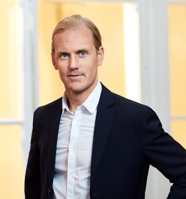 Mies mustassa takissa ja valkoisessa paidassa seisoo kirkkaiden ikkunoiden edessä.