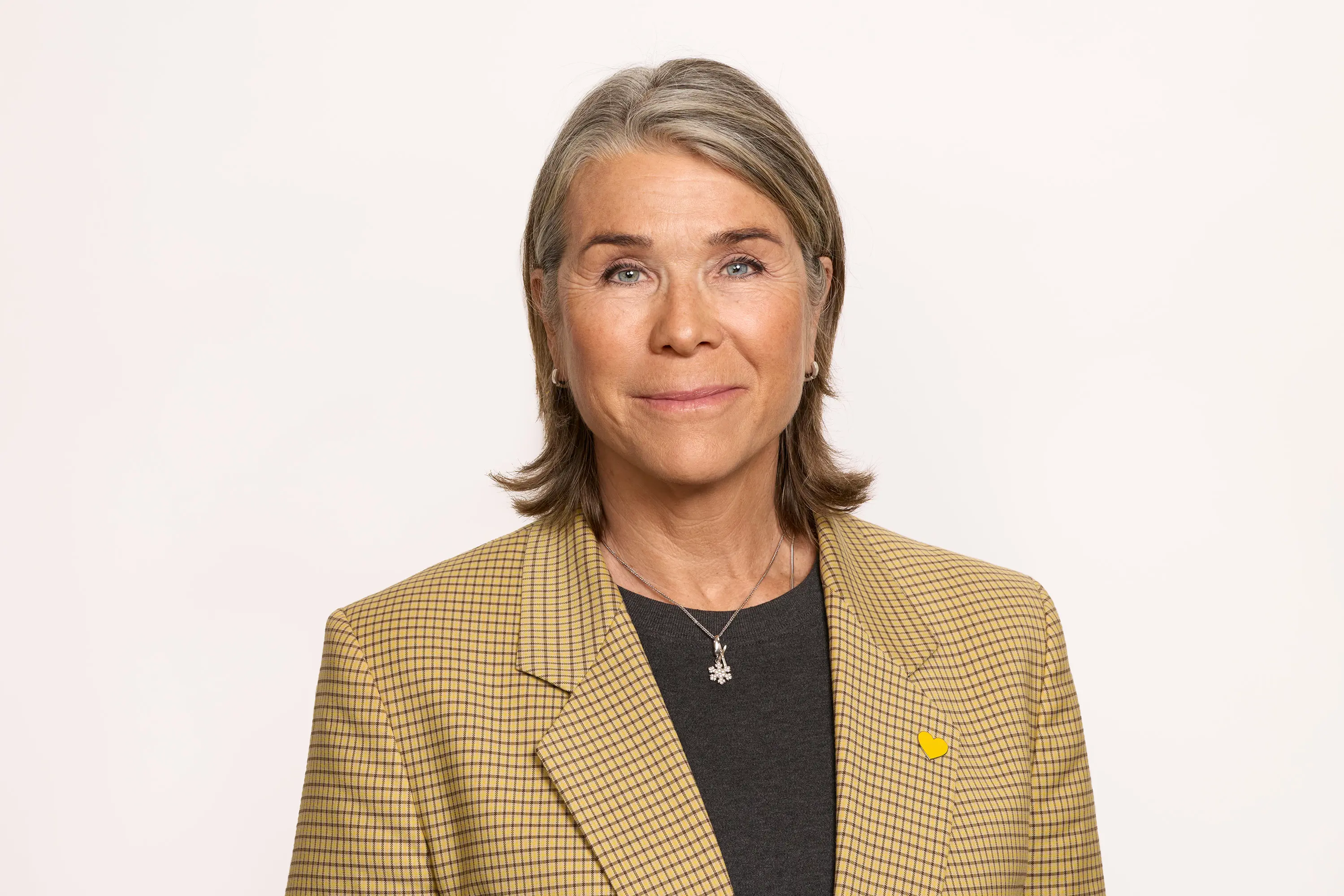 Kvinna med kort grått hår och brun- och svartmönstrad kavaj mot vit bakgrund, ser in i kameran med ett vänligt uttryck.
