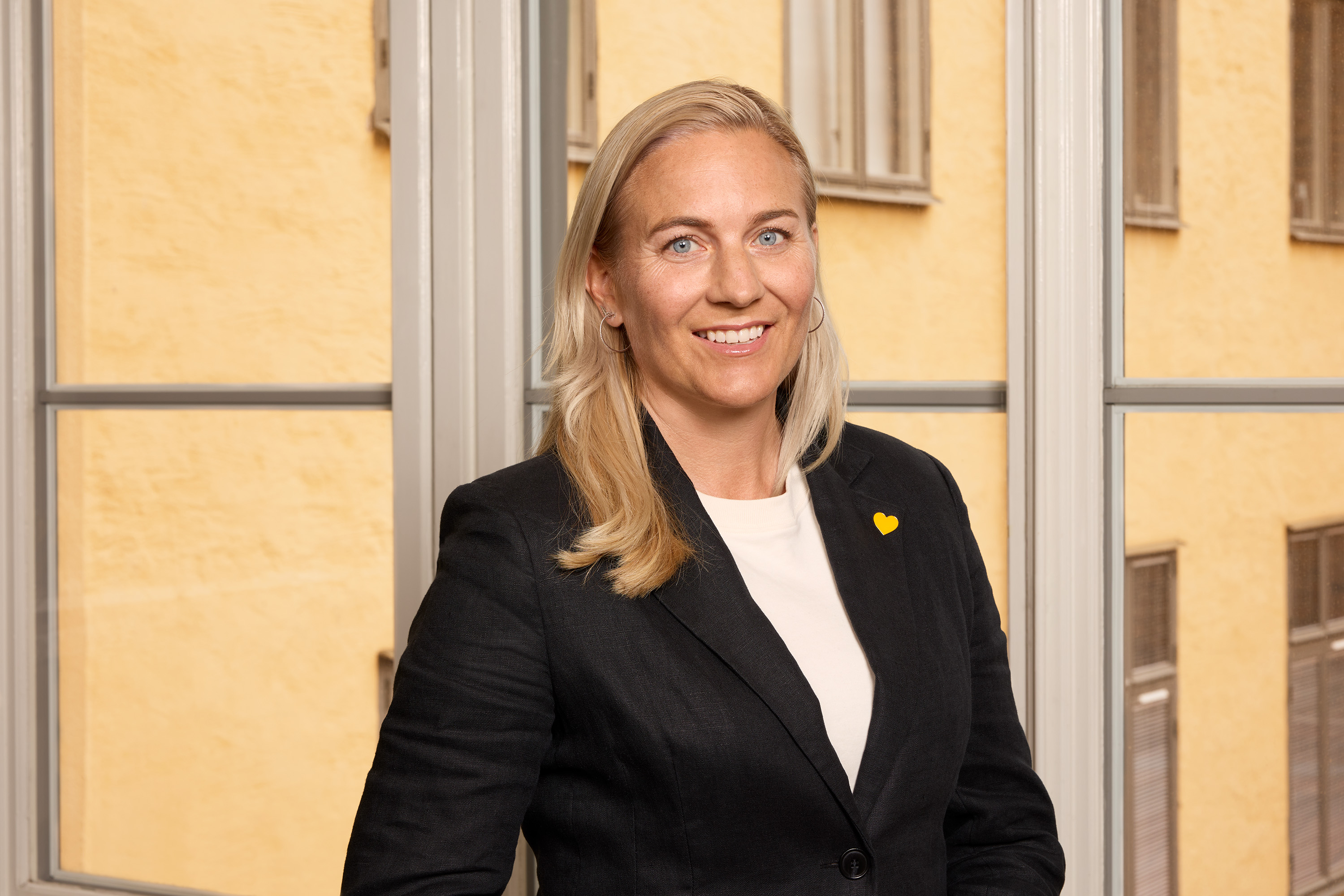 Nainen bisnespuvussa keltainen neula seisoo huoneessa, jossa on suuret ikkunat ja keltaiset seinät.