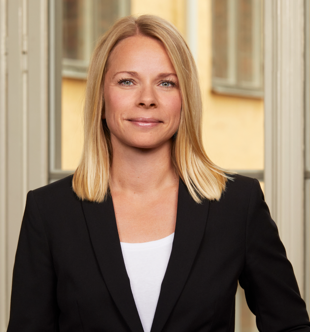 Nainen mustassa bleiserissä, jolla on pitkät vaaleat hiukset, seisoo ikkunan edessä.