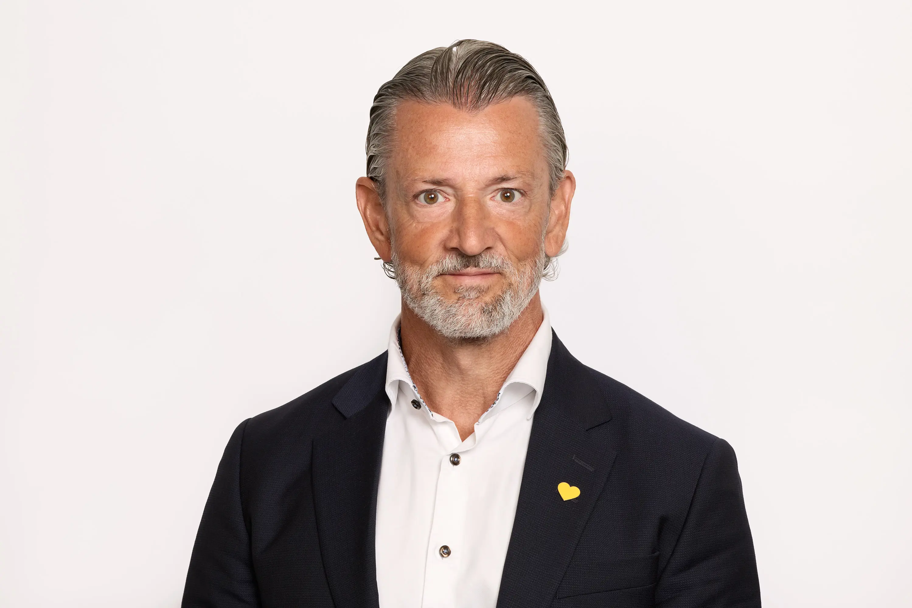 Man med grått hår och skägg i kostym och vit skjorta, neutral bakgrund.