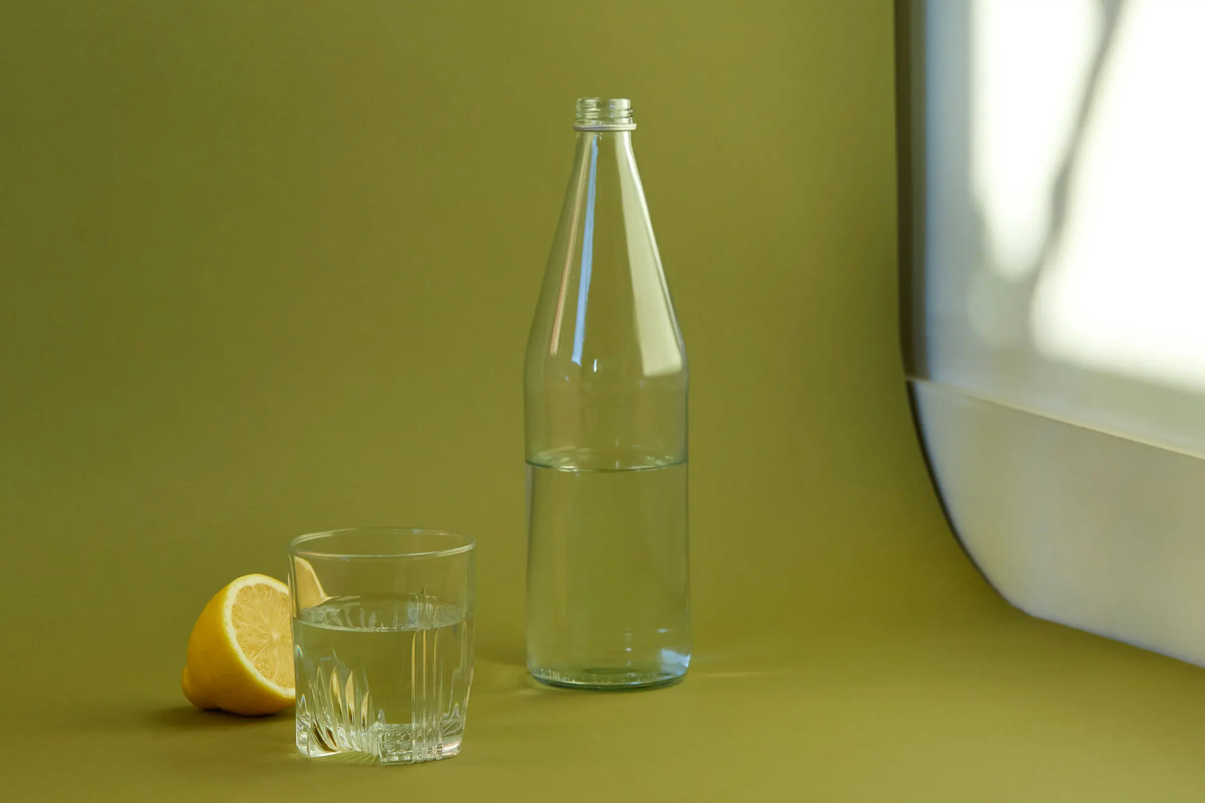 Glasflaska och glas med vatten bredvid en halv citron, placerade på ett grönt bord med solljus från ett fönster.