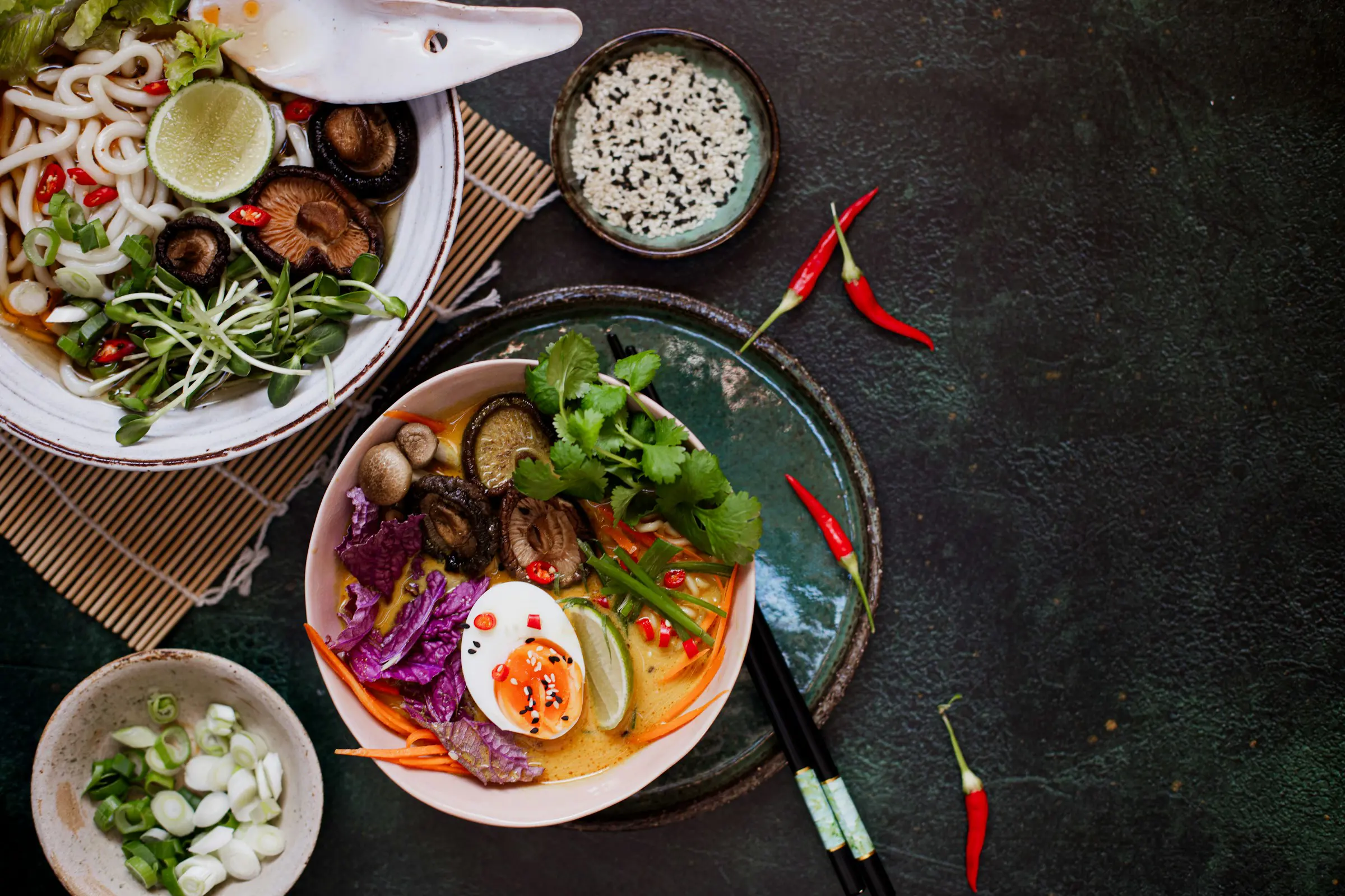 Färgglad skål med asiatisk nudelsoppa, innehållande nudlar, lime, svamp, koriander, rödkål och ett kokt ägg, omgiven av chili och salladslök på ett mörkt bord.