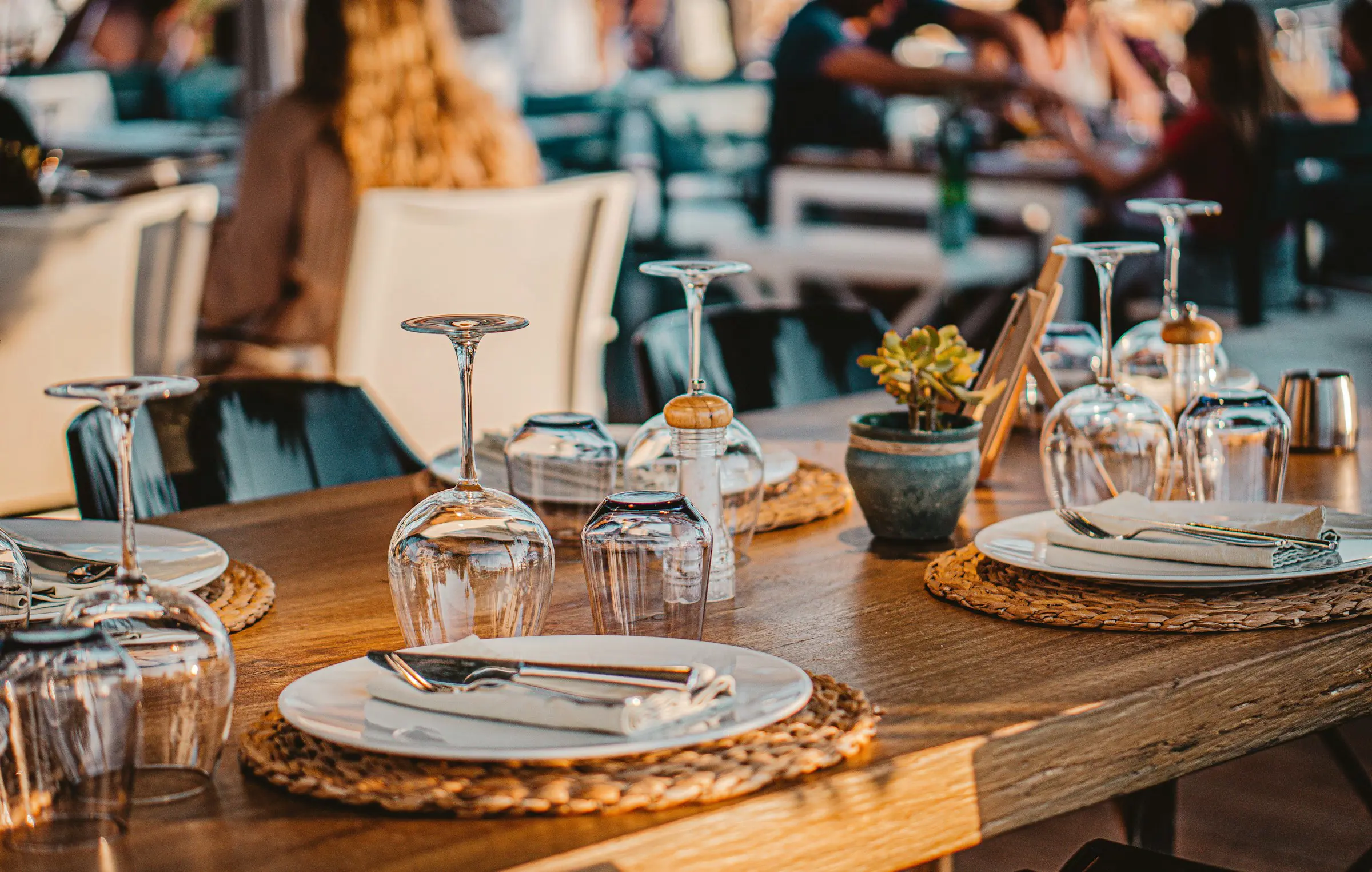 Dukat restaurangbord med glas och servetter i ett solbelyst rum, omgiven av människor i bakgrunden.