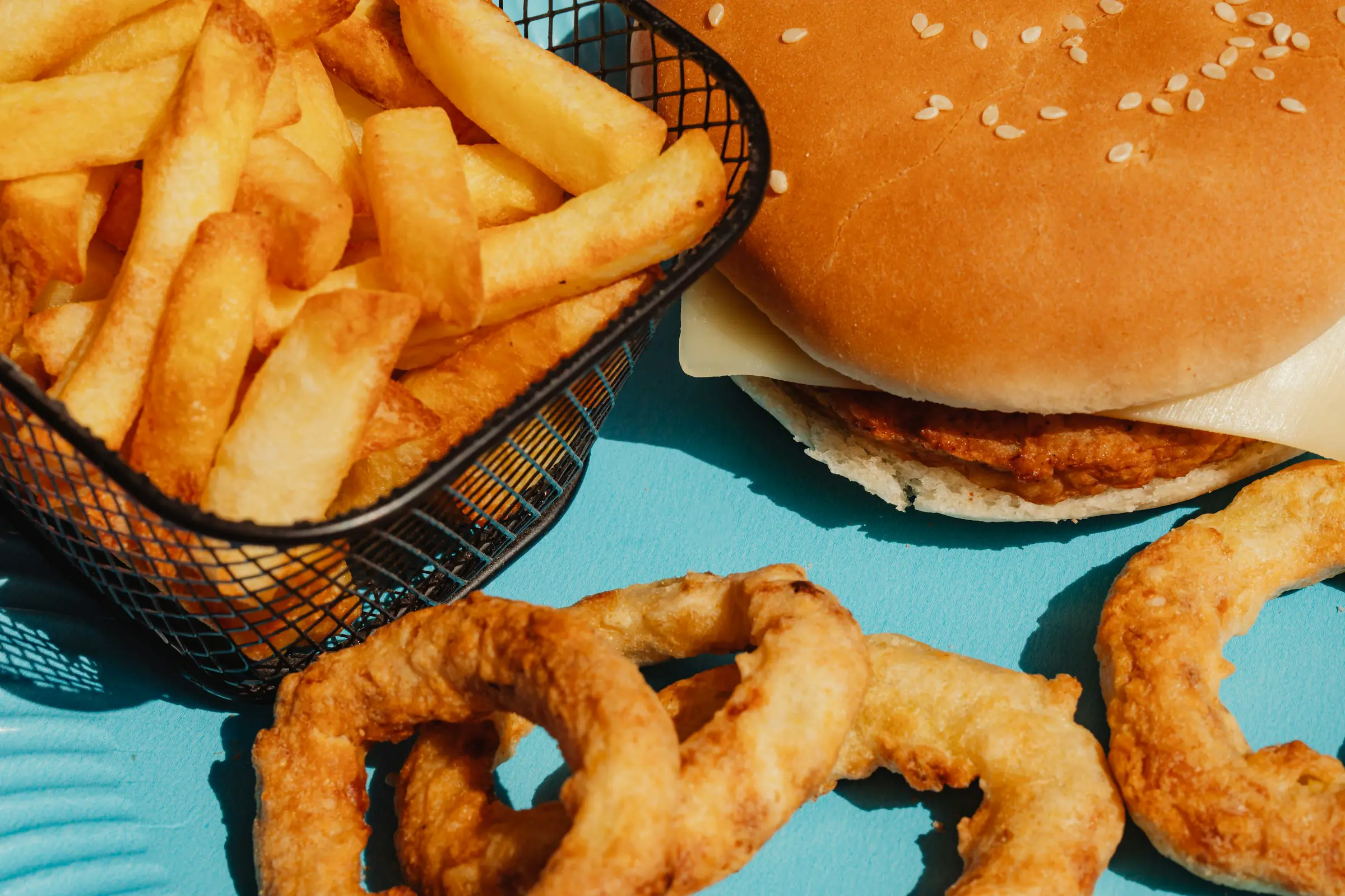 Hamburgare med ost, krispiga pommes frites i korg och friterade lökringar på blå bakgrund.